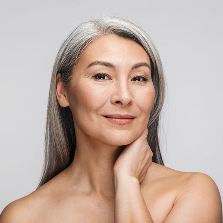 Beauty portrait of an attractive sensual mature topless woman with long gray hair standing isolated over gray background, posing, looking at camera