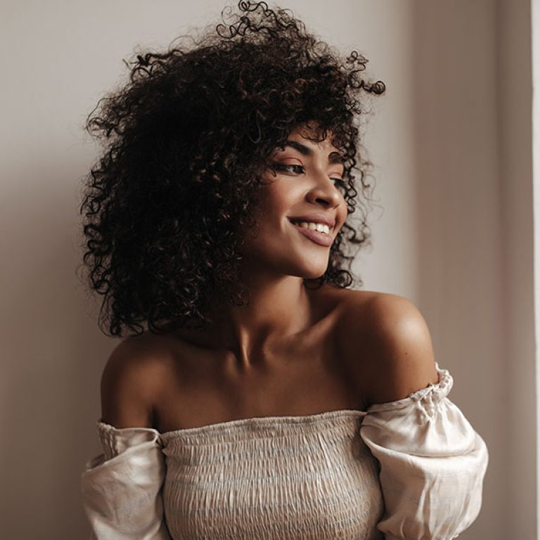Attractive dark-skinned woman leans on white wall. Pretty curly brunette lady in stylish top smiles sincerely.