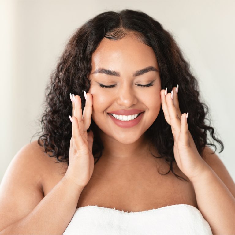 Happy oversize woman touching her face and smiling with closed eyes, making beauty treatments at home. Young body positive lady enjoying selfcare